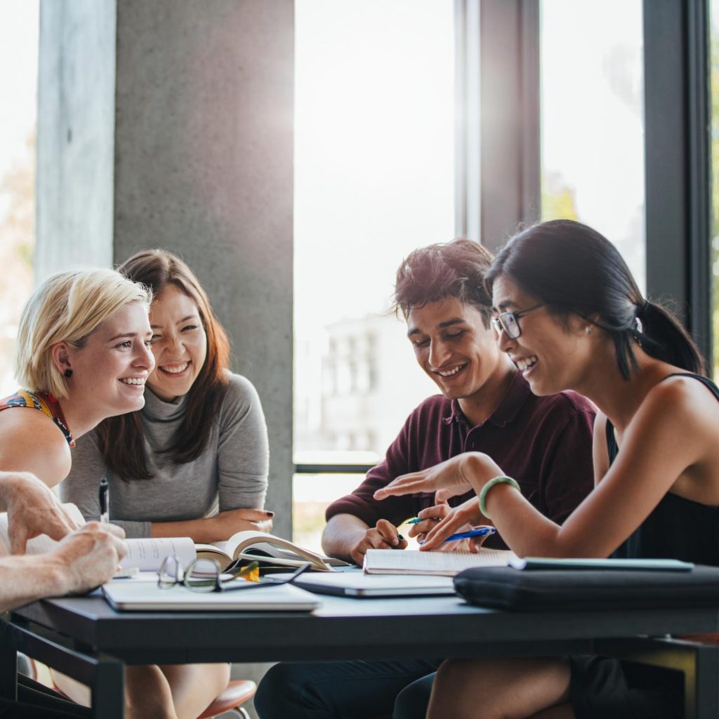 Four individuals engaged in a discussion around a table with notebooks and pens, in a room with large windows providing ample natural light.